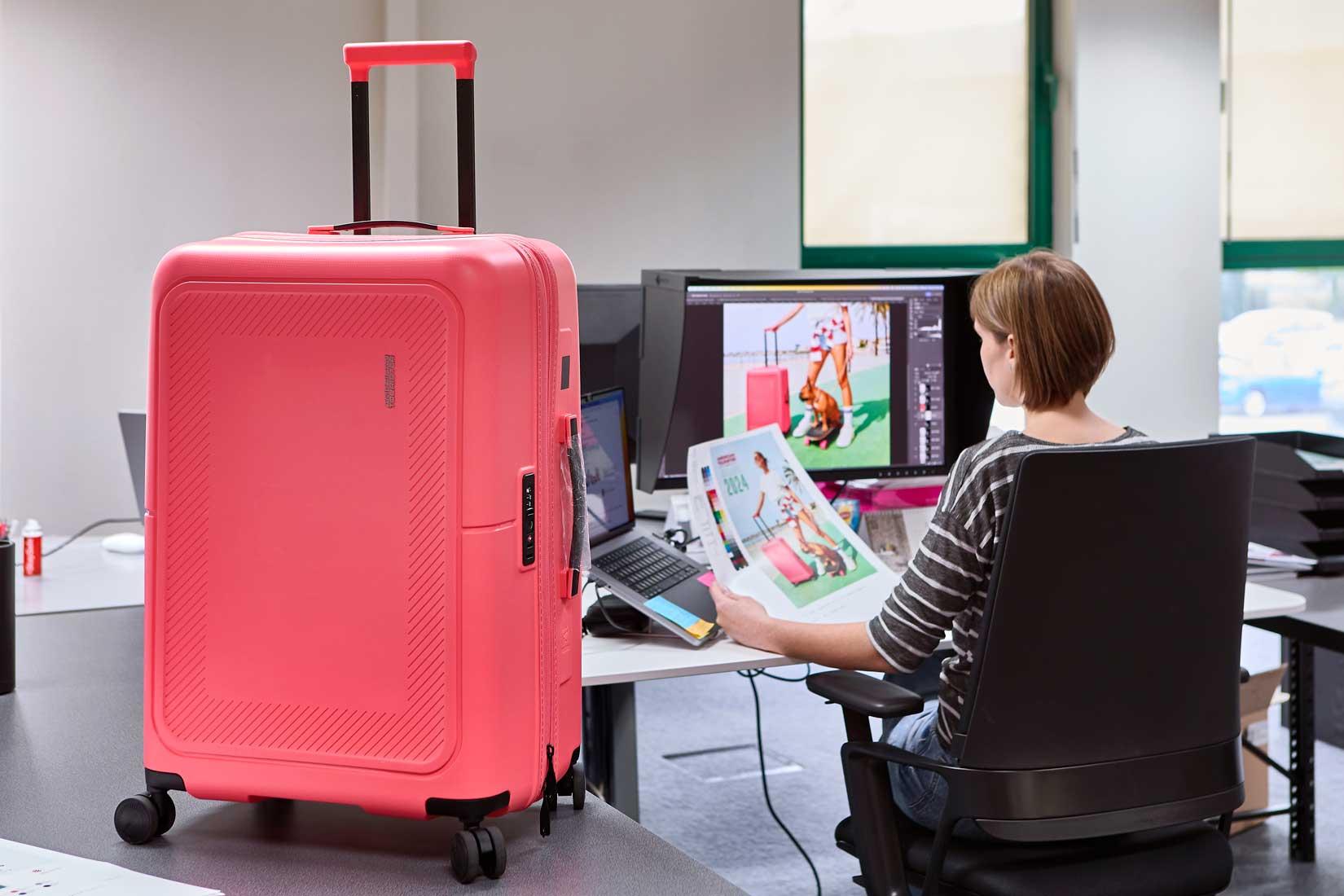 Roze American Tourister reiskoffer in een kantooromgeving, naast een designer die beeldmateriaal controleert op een computerscherm.