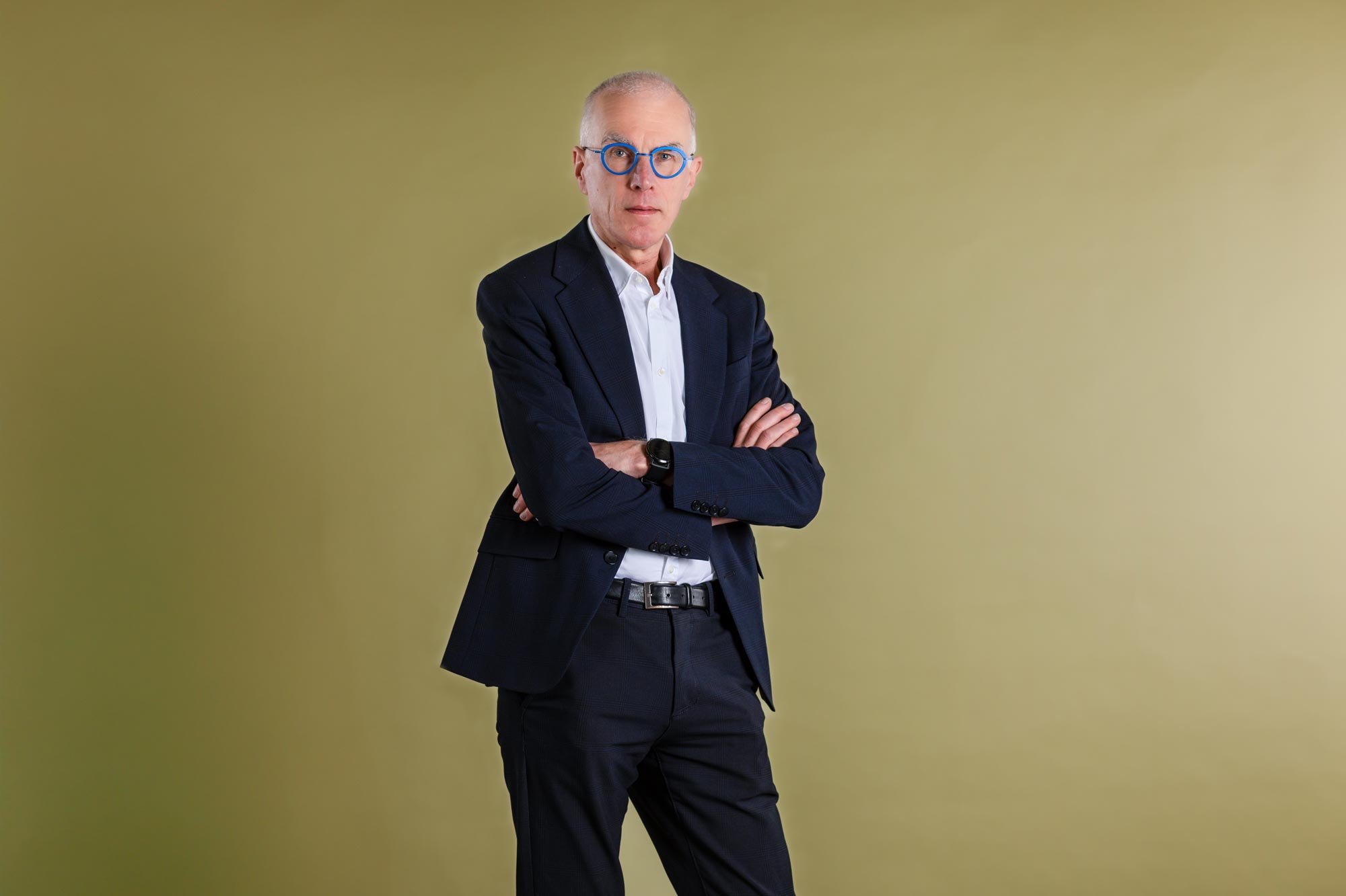Zakelijk portret van Jan Balliauw, staand met gekruiste armen, blauwe bril en donker pak tegen een effen groene achtergrond.