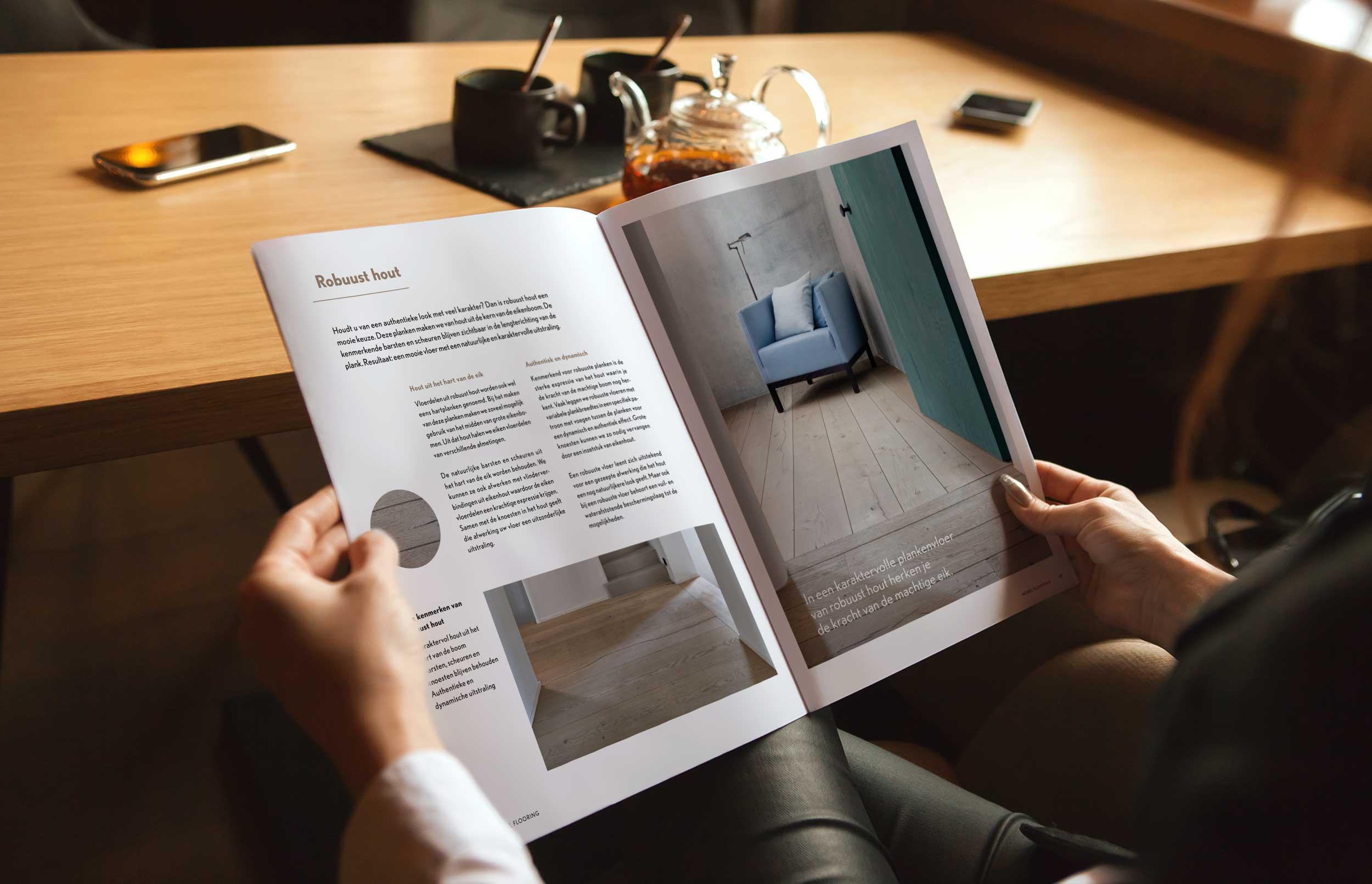 Persoon bladert door een brochure over houten vloeren aan een houten tafel, met foto’s van interieurtoepassingen en materiaalinformatie.