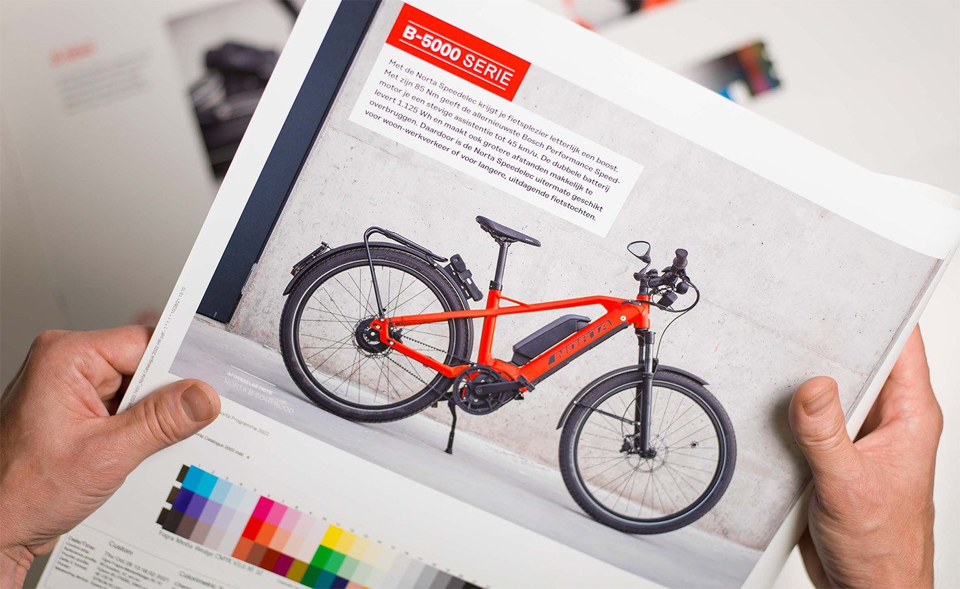 Handen die een drukproef van een Norta-catalogus vasthouden, met een foto van een feloranje elektrische fiets en kleurstalen onderaan het drukvel.