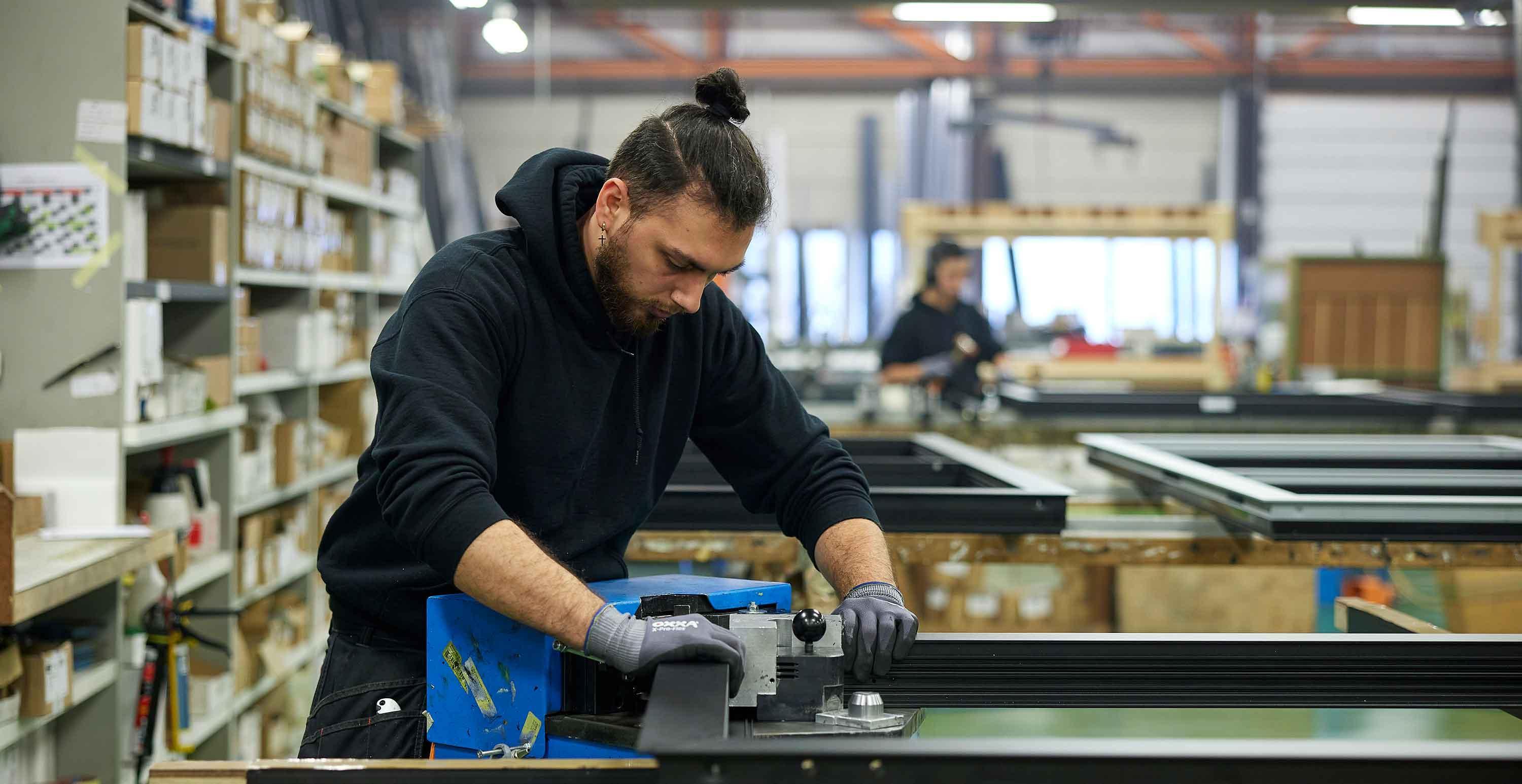 Medewerker werkt geconcentreerd aan een machine in een productiehal, omringd door metalen profielen en industriële apparatuur.