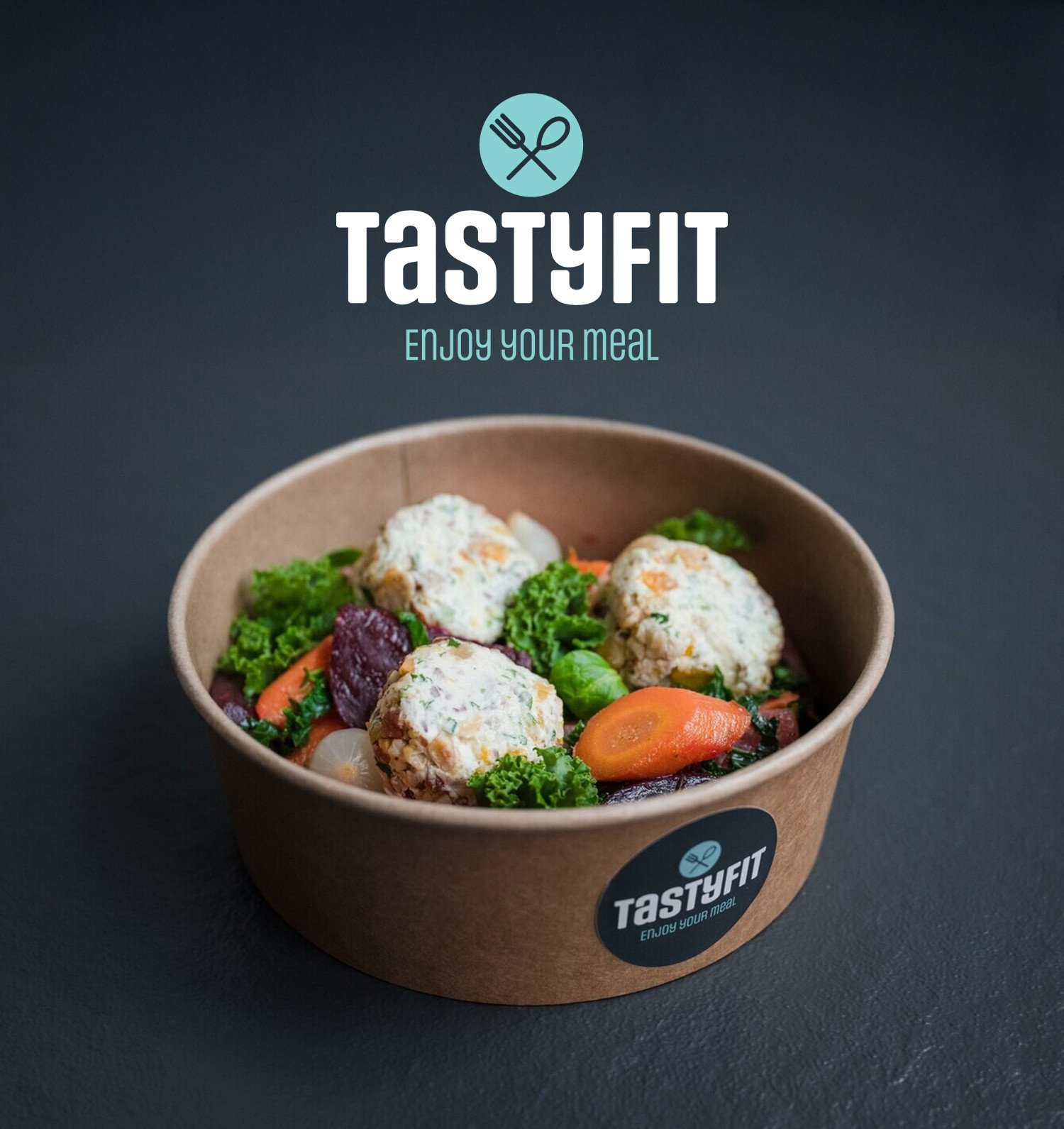 Een take away box gevuld met kleurrijke groenten en drie ronde vegetarische balletjes, geplaatst op een donkere ondergrond. Boven de kom staat het TastyFit-logo met de slogan ‘Enjoy your meal’.