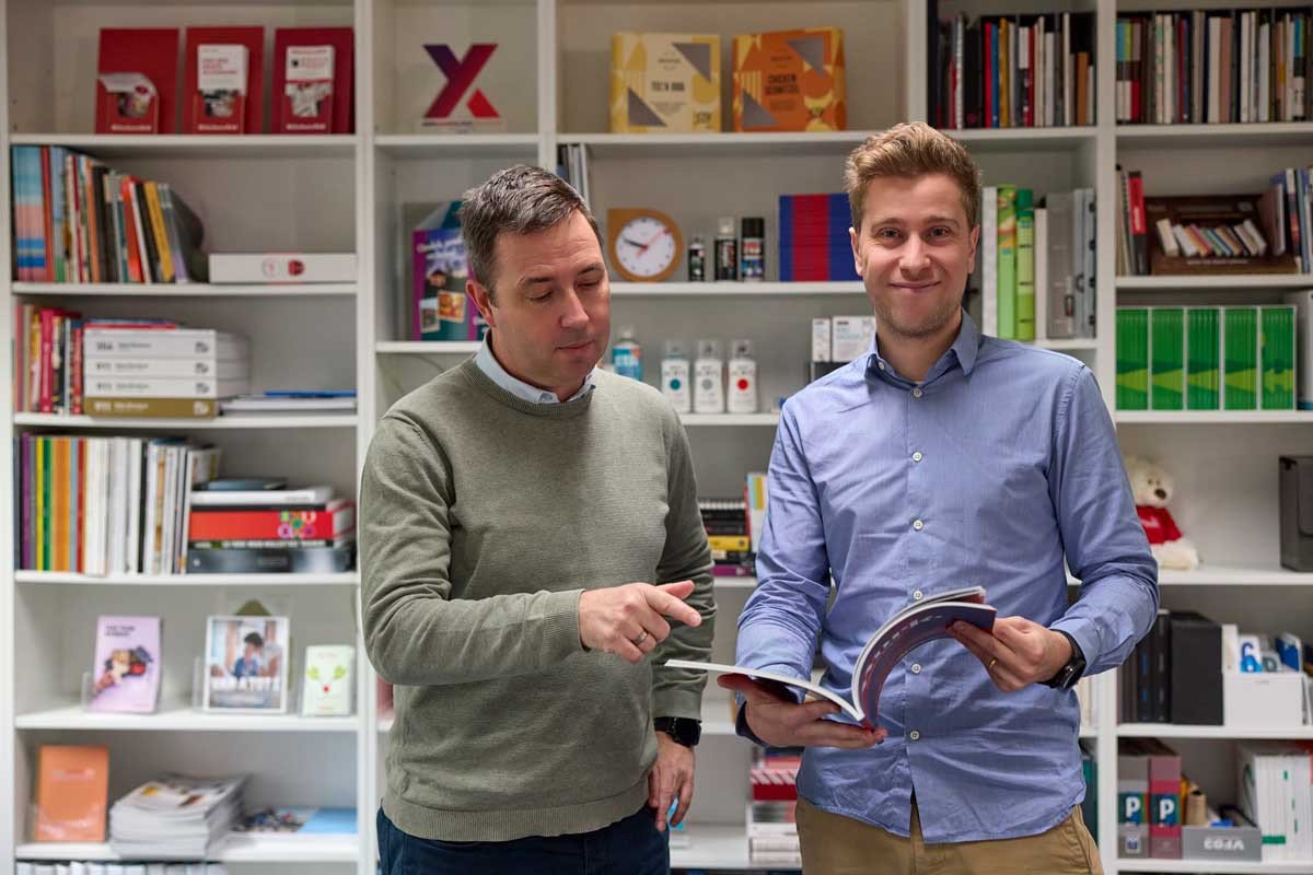 Twee collega’s bekijken samen een brochure in een kantooromgeving met boekenkast.